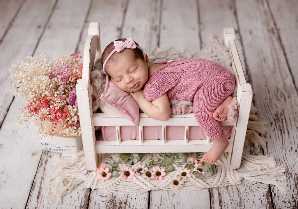 Sleeping newborn in a pink knit outfit on a miniature bed with flowers