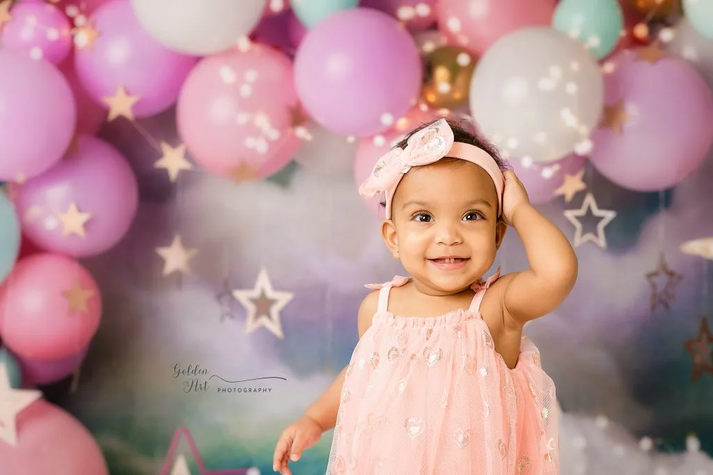 Smiling baby in a pink dress in front of a pastel balloon backdrop